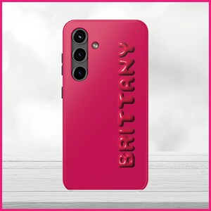 Peut inclure: Une coque de t&eacute;l&eacute;phone rose vif avec un lettrage blanc en relief qui &eacute;pelle "Brittany". La coque a un triple objectif d'appareil photo au dos.