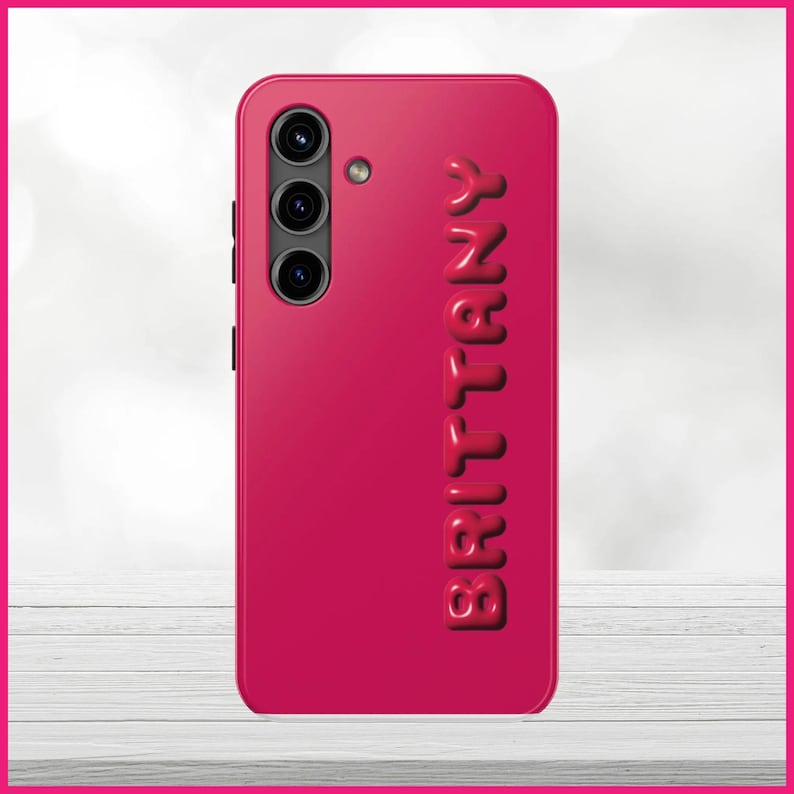 Peut inclure: Une coque de t&eacute;l&eacute;phone rose vif avec un lettrage blanc en relief qui &eacute;pelle "Brittany". La coque a un triple objectif d'appareil photo au dos.