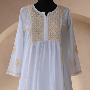 May include: White long-sleeve dress with intricate yellow embroidery around the neckline, sleeves, and bodice. The dress features a split neckline and a flowing, comfortable design. The embroidery includes floral and geometric patterns.