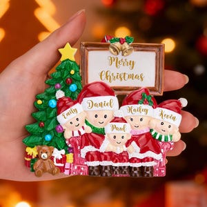 May include: A festive Christmas ornament depicting a family of five in Santa hats and red outfits. The ornament includes a Christmas tree, a teddy bear, and a sign with the words "Merry Christmas".