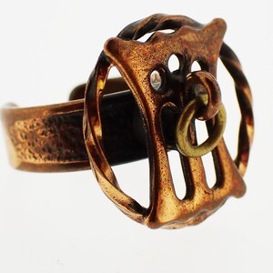 Peut inclure: Une bague en cuivre avec un design détaillé en forme de cercle avec un cercle plus petit suspendu en bas. La bague est en métal texturé et a un aspect rustique.