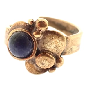 Op de afbeelding: Een goudkleurige ring met een donkerblauwe, ronde edelsteen. De ring heeft een organisch, getextureerd ontwerp met verhoogde, afgeronde details rond de centrale steen. De band is een eenvoudig, glad ontwerp.