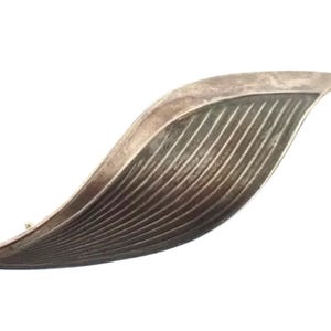 Peut inclure: Broche argentée en forme de feuille stylisée. La broche a un design incurvé et allongé avec un motif texturé et nervuré sur la partie inférieure. Le bord supérieur est lisse et arrondi.