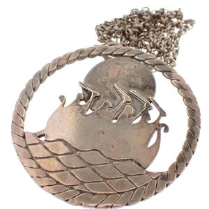 May include: A silver pendant necklace featuring a circular design with a ship sailing on the water. The pendant is surrounded by a rope-like border.