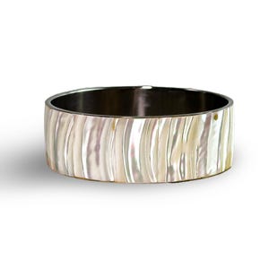 Puede incluir: Una pulsera rígida ancha con incrustaciones de nácar iridiscente y una textura vertical ondulada. La superficie exterior muestra un acabado brillante en blanco nacarado y crema, mientras que la banda interior es de un tono metálico oscuro.