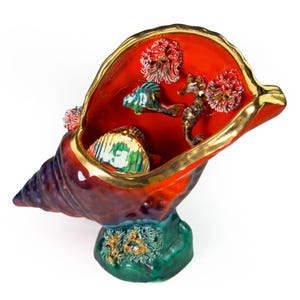 Peut inclure: Une sculpture en céramique en forme de coquillage avec un intérieur rouge vif et des bordures dorées. À l'intérieur, des créatures marines miniatures, dont un hippocampe, un poisson et du corail. La base est verte avec des accents dorés.