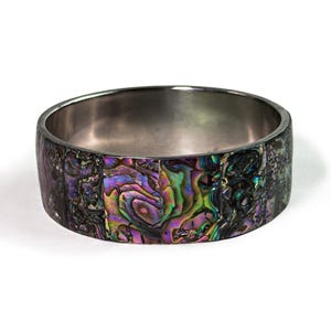 Puede incluir: Una pulsera rígida ancha que presenta un mosaico de segmentos de concha de abulón iridiscente. El exterior muestra una vibrante gama de tonos morados, verdes y azules con un brillo nacarado natural. El interior es de metal plateado liso y pulido.