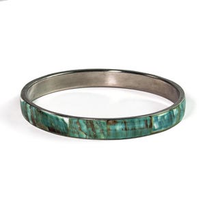 Könnte beinhalten: Ein kreisförmiger Armreif mit einem dunklen Metallband, das mit rechteckigen Segmenten aus poliertem blaugrünem und braunem Stein eingelegt ist. Die Außenfläche zeigt ein natürliches Marmormuster mit verschiedenen Grüntönen und dunkelbraunen Streifen.