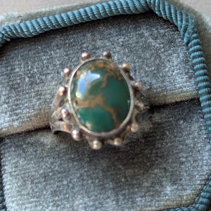 Op de afbeelding: Een zilveren ring met een ovale, groene steen met bruine adering. De ring heeft een decoratieve zetting met kleine, ronde accenten rond de steen. Gepresenteerd in een grijze fluwelen doos met een blauwgroene rand.