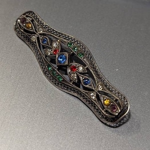 Puede incluir: Un broche de filigrana de plata con un diseño delicado que presenta piedras preciosas de colores. El broche está engastado con piedras azules, rojas, verdes y amarillas.
