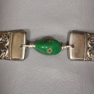 May include: A silver bracelet featuring a green and white bead. The bracelet is crafted from two silver sections with elaborate designs and a green bead with white floral details.