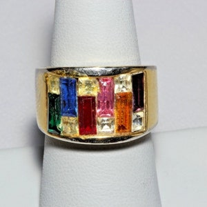 Puede incluir: Un anillo de oro con una banda de piedras rectangulares de colores. Las piedras están engastadas en una banda de plata y están dispuestas en fila. El anillo está en un expositor blanco.
