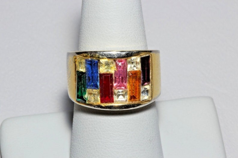 May include: A gold ring with a band of colorful rectangular stones. The stones are set in a silver band and are arranged in a row. The ring is on a white display stand.