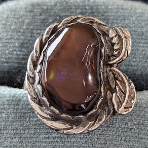 May include: A silver ring with a large, oval, brown gemstone. The ring has a braided border and leaf accents on the sides. The gemstone has a glossy, reflective surface.