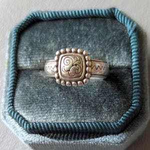 May include: Silver ring with a square, beaded center featuring a swirling design. The ring has a zig-zag pattern on the band and is displayed in a teal velvet ring box. A close-up shot highlights the ring's details.