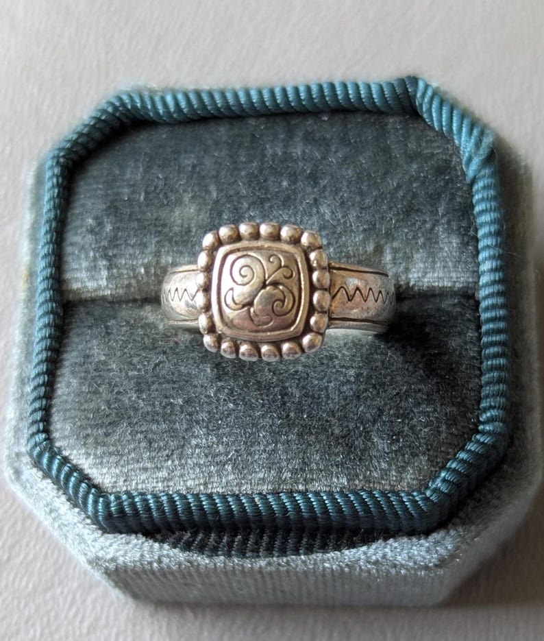 May include: Silver ring with a square, beaded center featuring a swirling design. The ring has a zig-zag pattern on the band and is displayed in a teal velvet ring box. A close-up shot highlights the ring's details.