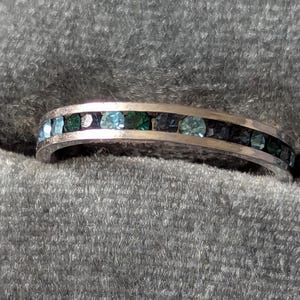 May include: A silver-toned metal band ring featuring a channel setting with a continuous row of small, round-cut gemstones in varying shades of teal, dark blue, and deep green, creating a gradient effect across the surface of the jewelry piece.