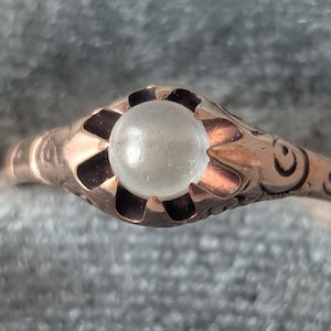 May include: A close-up of a vintage gold ring with a pearl-like, round, off-white stone. The ring has an ornate, floral-inspired design around the stone and along the band. The ring is set against a soft, gray background.