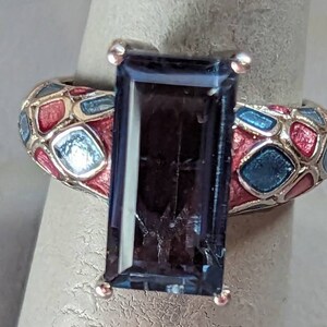 Könnte beinhalten: Ein silberner Ring mit einem großen, dunkelblauen rechteckigen Edelstein, der in einer Zargenfassung sitzt. Die Ringschiene ist mit einem Mosaik aus rotem und blauem Email verziert.