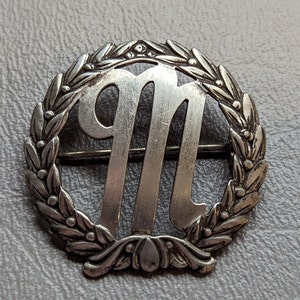 Może przedstawiać: Srebrna broszka z ozdobnym wzorem laurowym otaczającym literę "M".