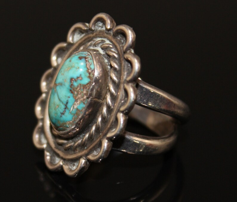 May include: A silver ring with a turquoise stone set in a detailed, scalloped design. The ring has a double band.