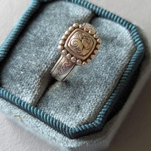 May include: A silver ring with a square, ornate face featuring a swirling design. The ring is set with small, round beads around the face and has a textured band. The ring is displayed in a teal velvet ring box.