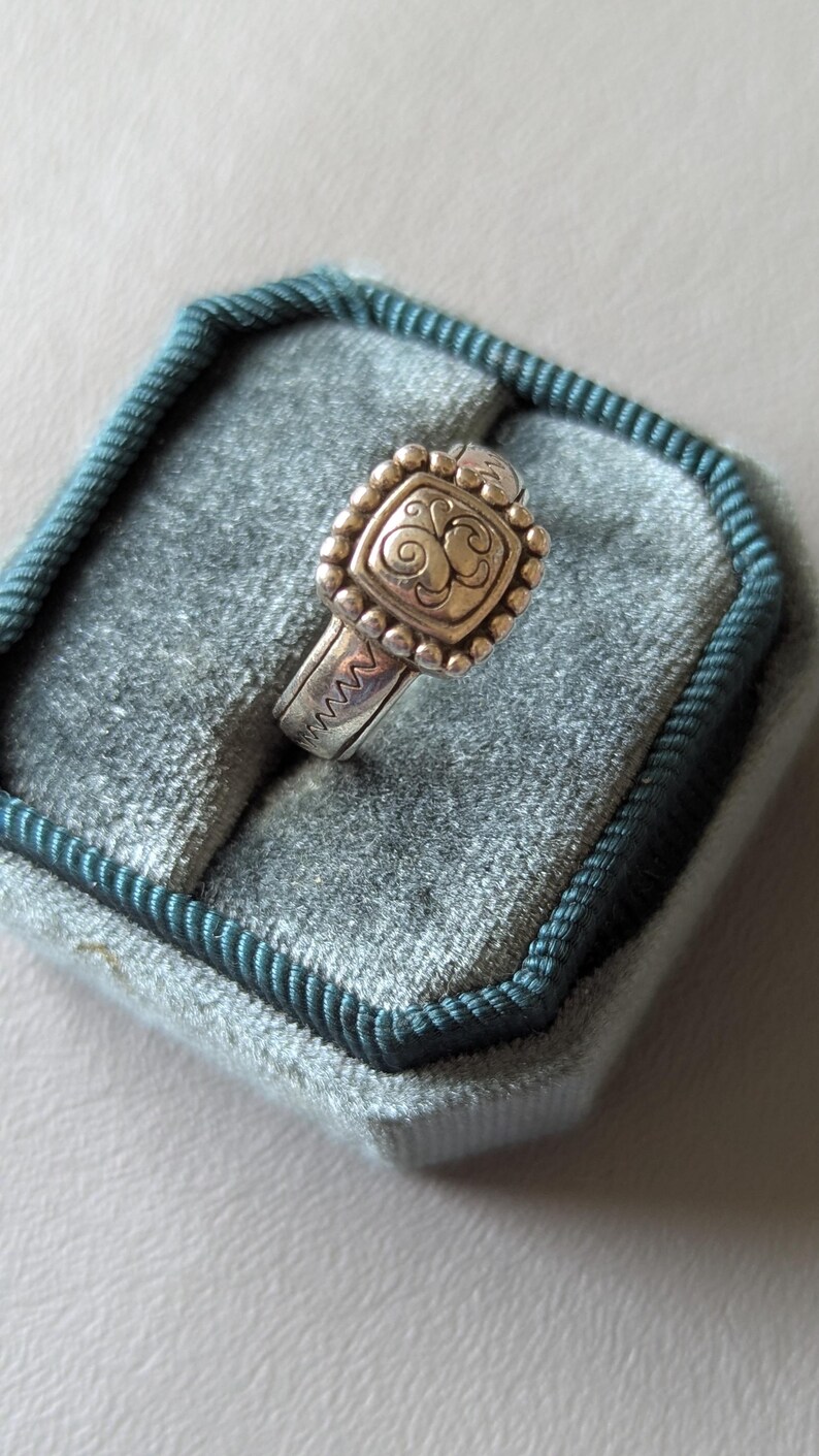 May include: A silver ring with a square, ornate face featuring a swirling design. The ring is set with small, round beads around the face and has a textured band. The ring is displayed in a teal velvet ring box.