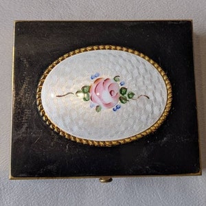 Op de afbeelding: Een zwart emaille compact met een gouden rand en een wit emaille ovaal met een roze roos erop geschilderd.