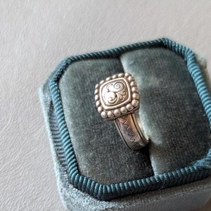 May include: A silver ring with a square, beaded top featuring a stylized letter "S" design. The ring is set on a teal velvet ring box with a dark teal border. The ring band has a zig-zag pattern.