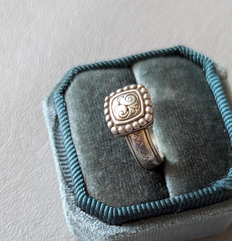 May include: A silver ring with a square, beaded top featuring a stylized letter "S" design. The ring is set on a teal velvet ring box with a dark teal border. The ring band has a zig-zag pattern.