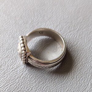 May include: A silver ring with a detailed design. The ring features a central circular element with an intricate pattern, surrounded by small beaded accents. The band has a textured, wavy pattern. The ring is shown on a light gray surface.