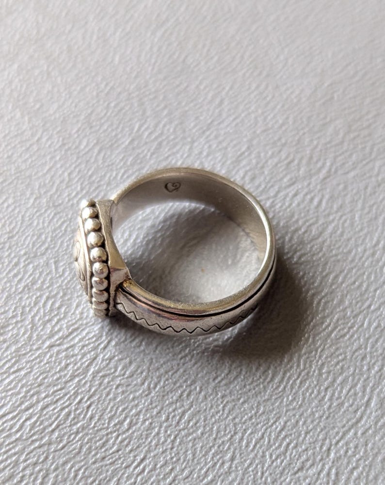 May include: A silver ring with a detailed design. The ring features a central circular element with an intricate pattern, surrounded by small beaded accents. The band has a textured, wavy pattern. The ring is shown on a light gray surface.