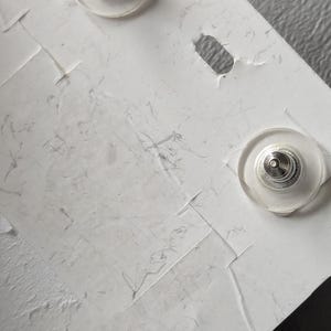 May include: Close-up of two silver-toned earring posts with clear, round backs, attached to a textured white card. The card has a small, oval-shaped hole and visible creases.