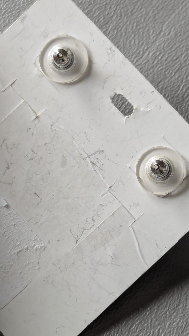 May include: Close-up of two silver-toned earring posts with clear, round backs, attached to a textured white card. The card has a small, oval-shaped hole and visible creases.
