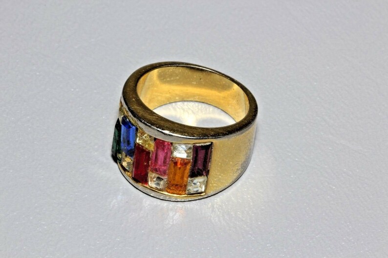 May include: A gold ring with a band of colorful rectangular stones. The stones are set in a silver band and are arranged in a row. The ring is a statement piece and would be a great addition to any jewelry collection.