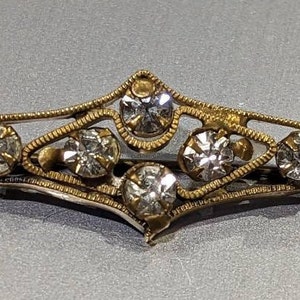 May include: A gold-toned brooch with a delicate filigree design and clear rhinestones.