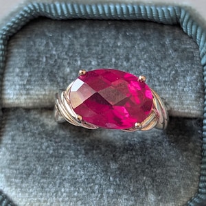 May include: A ring featuring a large, oval-cut, vibrant fuchsia-pink gemstone set in a polished silver-toned metal band. The stone is secured by four prongs, and the band displays a twisted, multi-strand design that tapers toward the center setting.