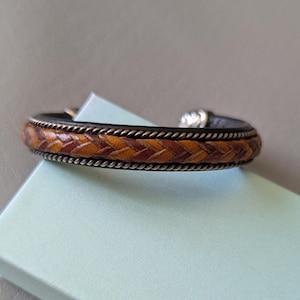 May include: A cuff bracelet featuring a central braided band of tan and dark brown leather. The leather braid is flanked by two thin, twisted metallic silver-toned wires. The bracelet has a dark brown leather base and a metallic silver-toned clasp.