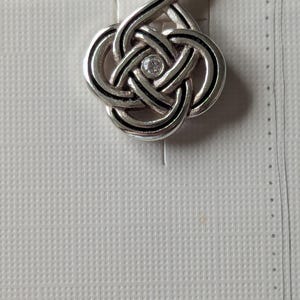 May include: Silver-toned pendant with a Celtic knot design, featuring a central clear gemstone. The pendant hangs from a loop and is displayed on a textured white card. The word "Brighton" is partially visible.