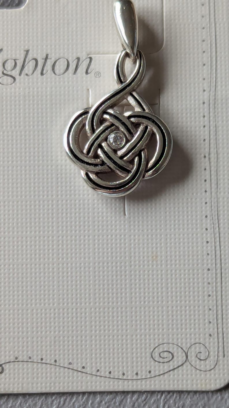 May include: Silver-toned pendant with a Celtic knot design, featuring a central clear gemstone. The pendant hangs from a loop and is displayed on a textured white card. The word "Brighton" is partially visible.