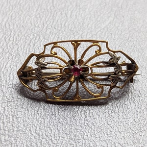 May include: A gold-toned brooch with a delicate filigree design and a single red gemstone in the center.