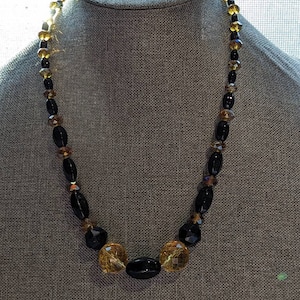 May include: A black and gold beaded necklace with oval-shaped beads. The necklace is displayed on a gray mannequin.