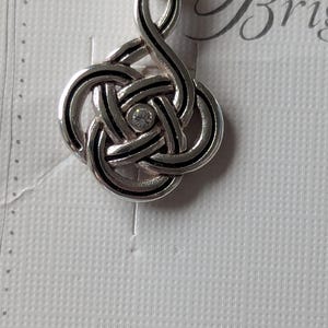 May include: A silver-toned pendant with a Celtic knot design. The knot is accented with black lines and a small, clear gemstone in the center. The pendant hangs from a silver bail. The background is a textured white surface.