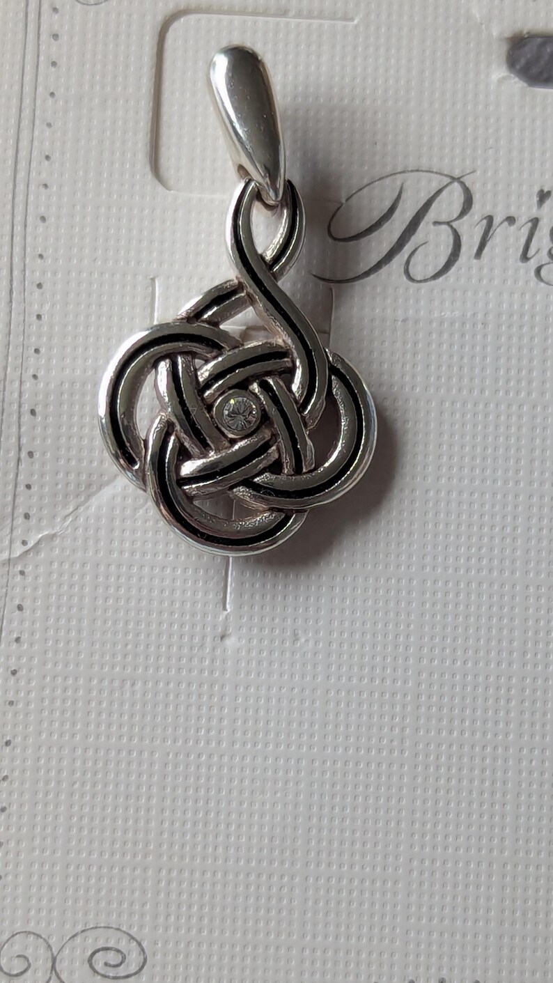 May include: A silver-toned pendant with a Celtic knot design. The knot is accented with black lines and a small, clear gemstone in the center. The pendant hangs from a silver bail. The background is a textured white surface.