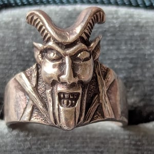 May include: A silver-toned ring featuring a detailed depiction of a devil's face. The ring showcases a horned head with a grimacing expression, open mouth, and pointed ears. The design is intricate, with visible facial features and textured details.