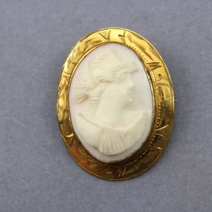Può includere: Spilla cammeo ovale dorata con profilo in rilievo bianco di donna. Il cammeo è incastonato in una cornice dorata decorativa. Un gioiello classico.