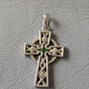 May include: A silver-toned Celtic cross pendant featuring an intricate knotwork design. The center of the cross is accented with a small, round green gemstone, while four clear, sparkling stones are embedded within the arms of the cross.