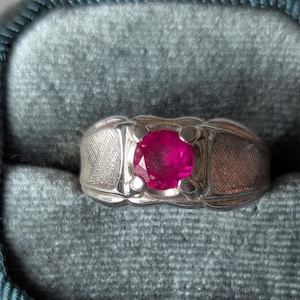 May include: A silver ring with a large, round, vibrant red gemstone. The ring has a textured band and is set against a teal velvet background, suggesting a jewelry display.