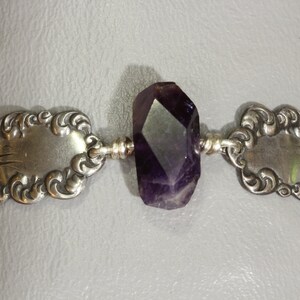 May include: A silver bracelet with a purple amethyst gemstone. The bracelet is made from a vintage spoon with ornate detailing.