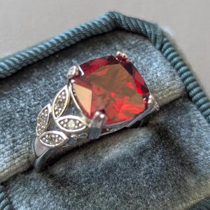 May include: A silver-toned ring featuring a large, square-cut, deep red gemstone as the centrepiece. The band is designed with a decorative leaf-shaped motif on the sides, which is accented with small, clear, sparkling stones set into the metal.
